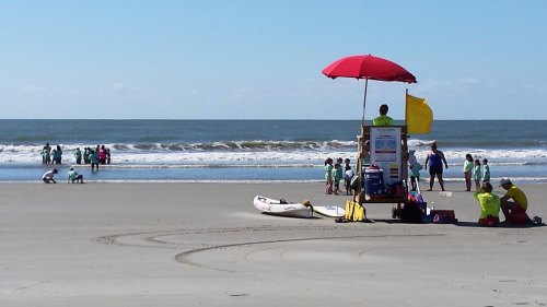 Lifeguard tower with red umbrella on a beach, people playing in the water, and a surfboard nearby.