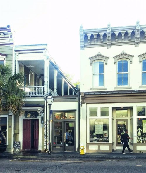A street view featuring historic buildings with shops, a person walking, and palm trees in the background.