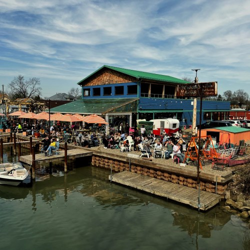 A bustling waterfront scene with a colorful restaurant, outdoor seating, and boats docked along the shore.