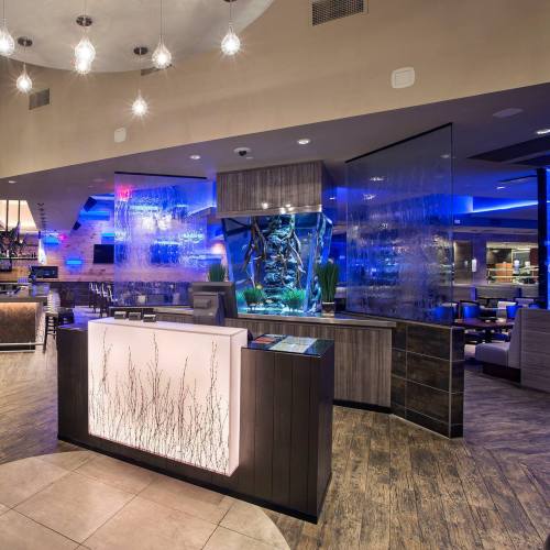 Modern restaurant interior featuring a sleek reception desk, illuminated water feature, and blue ambient lighting.