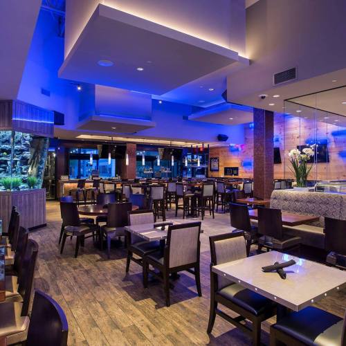 Modern restaurant interior with blue lighting, wooden floors, and empty tables and chairs.