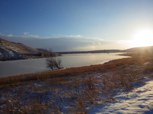 A serene winter landscape featuring a frozen lake, grassy banks, and a bright sun setting behind distant hills.
