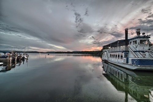 A serene lake at sunset with a paddleboat and moored boats reflecting on the water under a cloudy sky.