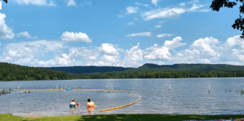 Two people wade in a lake surrounded by green hills and a blue sky with fluffy clouds. A swimming area is marked off.
