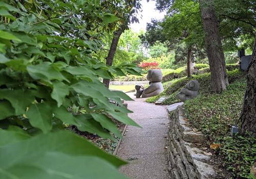 A serene garden path lined with greenery, featuring stone sculptures and trees in a peaceful setting.