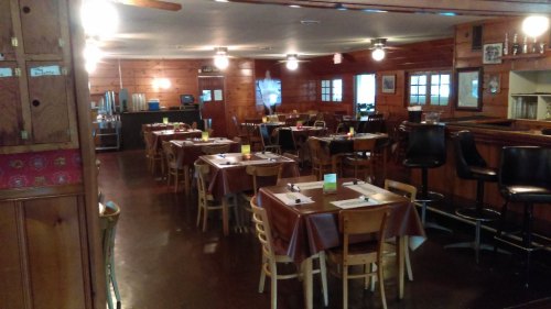Interior of a rustic restaurant with wooden walls, tables set for dining, and a bar area in the background.