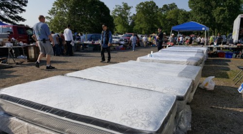 A row of new mattresses for sale at an outdoor market, with people browsing and tents in the background.