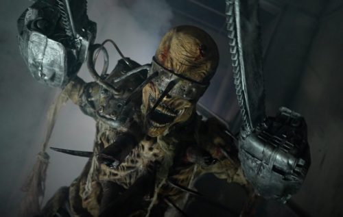 A menacing, skeletal creature with mechanical arms and a grotesque face, surrounded by smoke in a dimly lit environment.