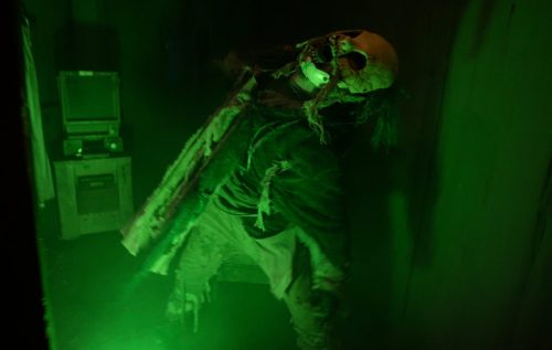 A creepy figure in a mask and tattered clothing stands in a dimly lit, green-hued room, creating a spooky atmosphere.