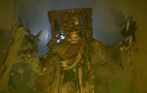 A creepy figure in tattered clothing and a cage-like mask stands in a dimly lit, eerie environment.