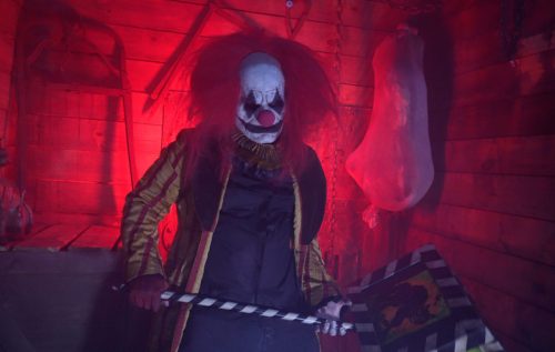 A sinister clown with a red wig and painted face stands in a dimly lit room, holding a striped weapon.
