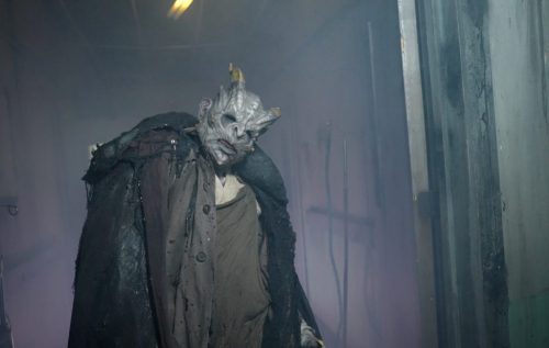 A creepy figure in a dark cloak with a monstrous face walks through a foggy, dimly lit corridor.