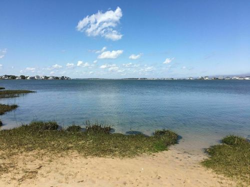 A serene coastal view featuring calm waters, sandy shore, and distant houses under a clear blue sky with scattered clouds.