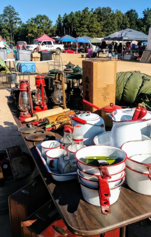 A table filled with vintage kitchenware, lanterns, and other items at an outdoor market with tents in the background.