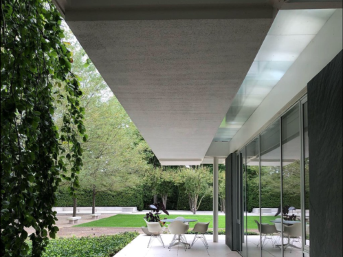 A modern outdoor space with a sleek roof, glass walls, and white chairs surrounded by greenery and a manicured lawn.