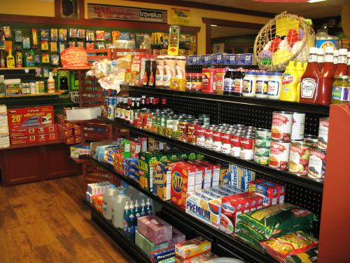 A well-stocked convenience store aisle featuring various canned goods, snacks, and condiments on black shelves.