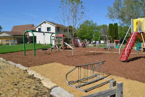 A sunny playground with swings, a slide, and a play structure, surrounded by grass and trees.