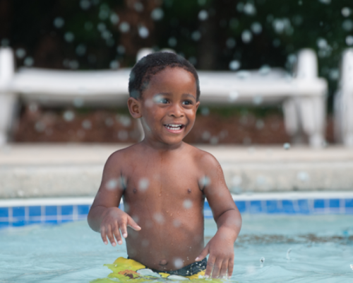 A smiling child stands in a pool, surrounded by splashing water droplets.