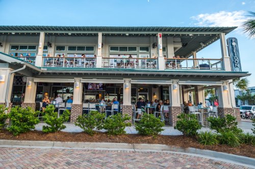 A lively restaurant with outdoor seating, bustling with patrons, surrounded by greenery and a clear blue sky.