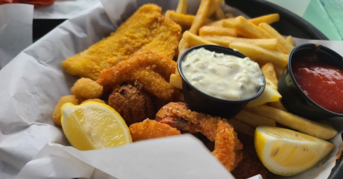 A plate of fried seafood including fish, shrimp, and fries, served with lemon wedges and dipping sauces.