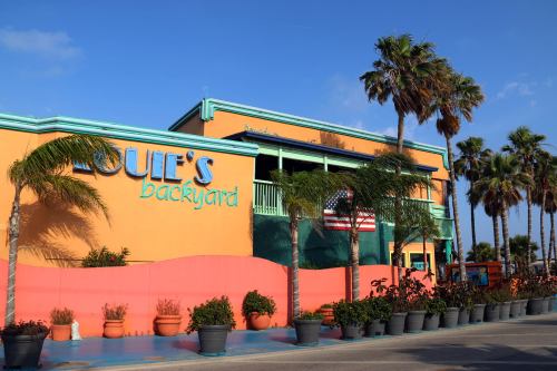 Colorful building with "Louie's Backyard" sign, palm trees, and an American flag, set against a clear blue sky.