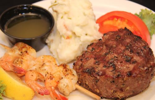 A plate featuring a grilled beef patty, shrimp skewers, mashed potatoes, a slice of tomato, and a lemon wedge.