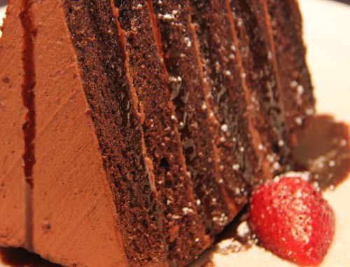 A slice of rich chocolate cake with layers, drizzled with chocolate sauce, and garnished with a fresh strawberry.