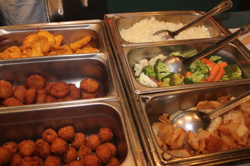 A buffet spread featuring fried shrimp, rice, mixed vegetables, and golden-brown fritters in metal serving trays.