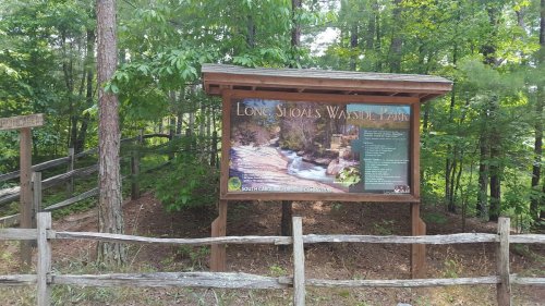 Sign for Long Shoals Wayside Park surrounded by trees, with a wooden fence in the foreground.