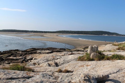 A scenic view of a beach with sandy shores, calm waters, and distant hills under a clear blue sky.