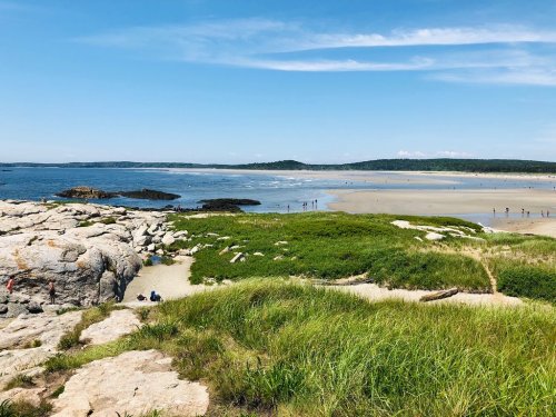 A scenic beach view with rocky shores, green grass, and people enjoying the sunny day under a clear blue sky.