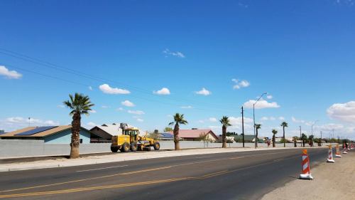 Construction equipment on a road lined with palm trees and houses under a clear blue sky with scattered clouds.