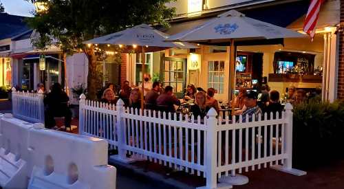 Outdoor dining scene at a restaurant with patrons seated under umbrellas, surrounded by a white picket fence.