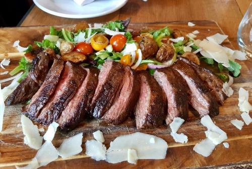 Sliced steak on a wooden board, surrounded by salad, cherry tomatoes, roasted potatoes, and shaved cheese.