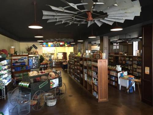 Interior of a market with wooden shelves, various products, and a vintage windmill ceiling fan. Brightly lit and inviting.