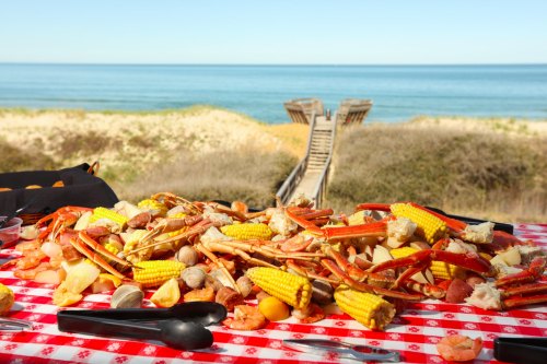 A seafood feast with crabs, shrimp, and corn on a red checkered tablecloth, overlooking a beach and ocean.