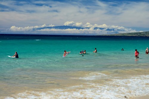 People enjoying a sunny day at the beach, swimming and surfing in clear turquoise waters with clouds in the background.
