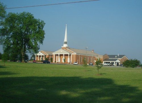 A brick church with a tall steeple, surrounded by green grass and trees under a clear blue sky.