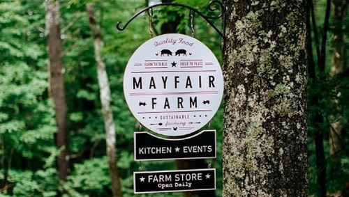 Sign for Mayfair Farm, featuring "Quality Food," with sections for Kitchen, Events, and Farm Store, surrounded by trees.