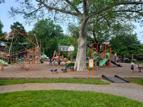 A playground with climbing structures, slides, and swings, surrounded by trees and children playing.