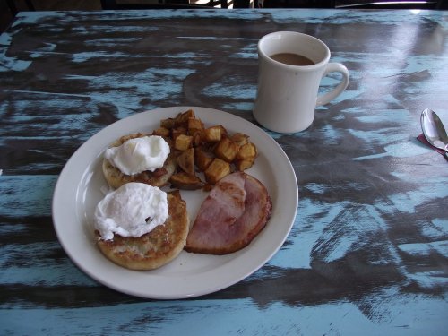 A plate of breakfast with poached eggs on English muffins, ham, roasted potatoes, and a cup of coffee on a blue table.