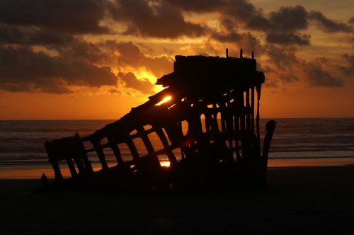 Silhouette of a shipwreck against a vibrant sunset over the ocean, with clouds in the sky.