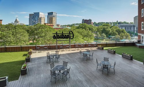 A spacious outdoor terrace with tables and chairs, overlooking a green park and city skyline under a clear blue sky.