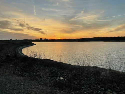 A serene sunset over a calm lake, with a curved shoreline and wispy clouds in the sky.