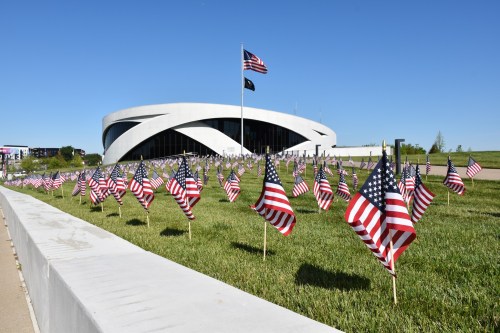 A field of small American flags in front of a modern building under a clear blue sky.