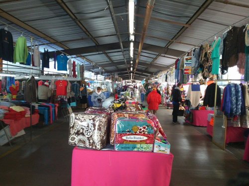A bustling indoor market with colorful clothing and blankets displayed on tables, shoppers browsing the aisles.