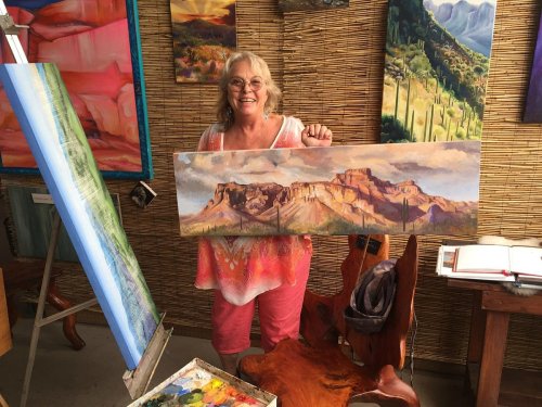 A smiling woman holds a landscape painting in an art studio, surrounded by colorful artworks and painting supplies.