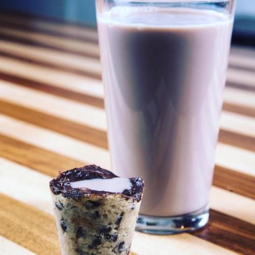 A chocolate chip cookie shot glass filled with milk next to a tall glass of chocolate milk on a wooden table.
