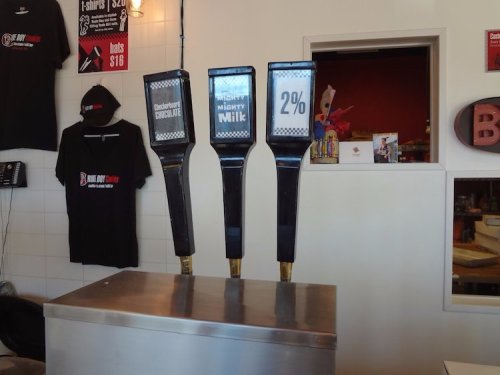 Three beverage taps labeled "Chocolate Milk," "Mighty Milk," and "2%," with a counter and shirts hanging in the background.