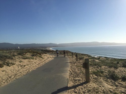 Two cyclists ride along a coastal path with sandy dunes and a view of the ocean under a clear blue sky.